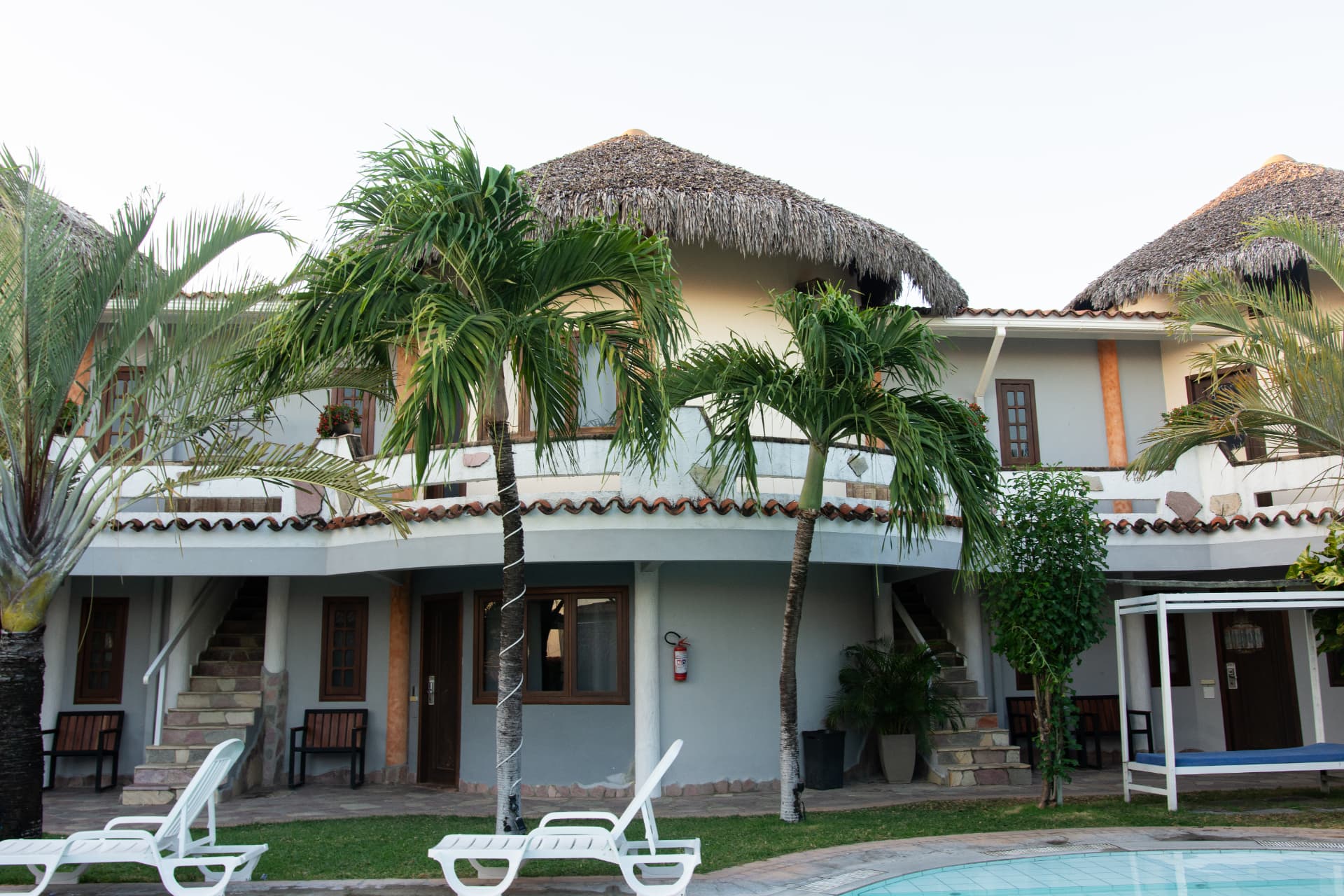 Fachada do hotel com telhados de palha e coqueiros vista da piscina